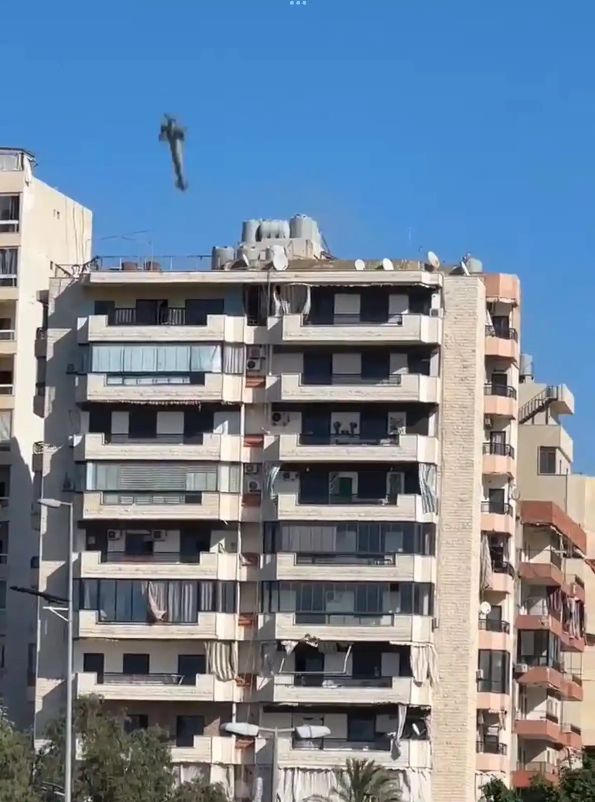 A blurry, distant figure appears to be floating or falling in the clear blue sky above a multi-story apartment building with…