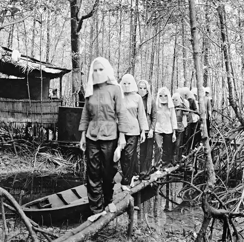 A black-and-white photo shows a line of people walking barefoot across a wooden log bridge in a swampy forest. Each person…