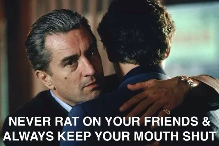 A still from a movie shows a man with graying hair and a serious expression embracing another man from behind. His hand…