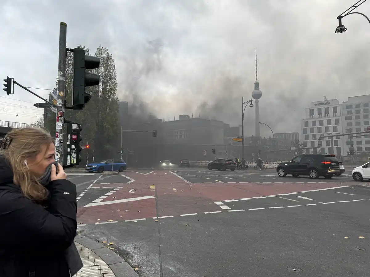 A woman in a black jacket stands at a Berlin street corner, covering her nose and mouth with a scarf as thick, dark smoke…