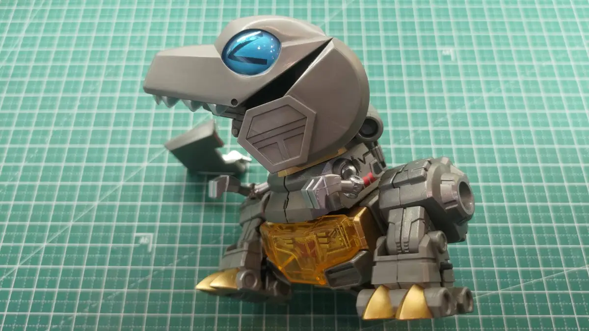 A small, grey robot dinosaur toy with a large blue eye and gold-colored feet sits on a green cutting mat. Its body is mostly…