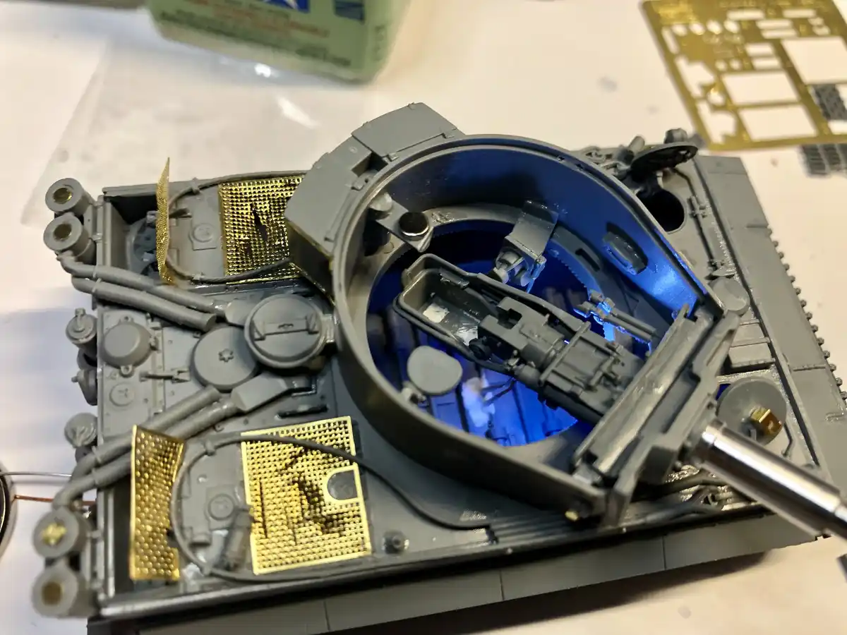 A close-up photo of an open, partially assembled grey plastic model tank with intricate internal parts, gold-colored metal…