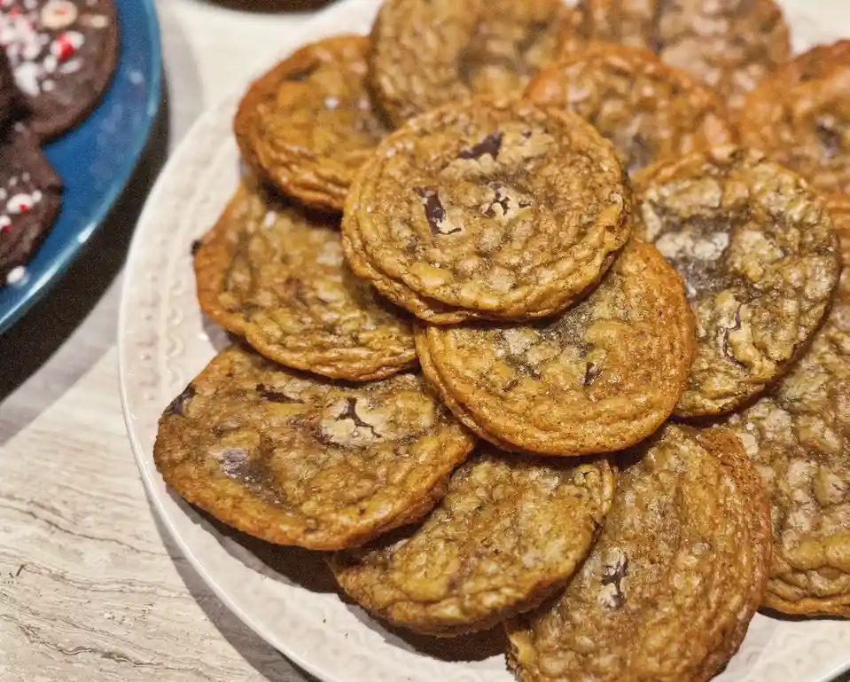 A close-up photo shows a white plate piled high with golden-brown chocolate chip cookies, some with visible chunks of…
