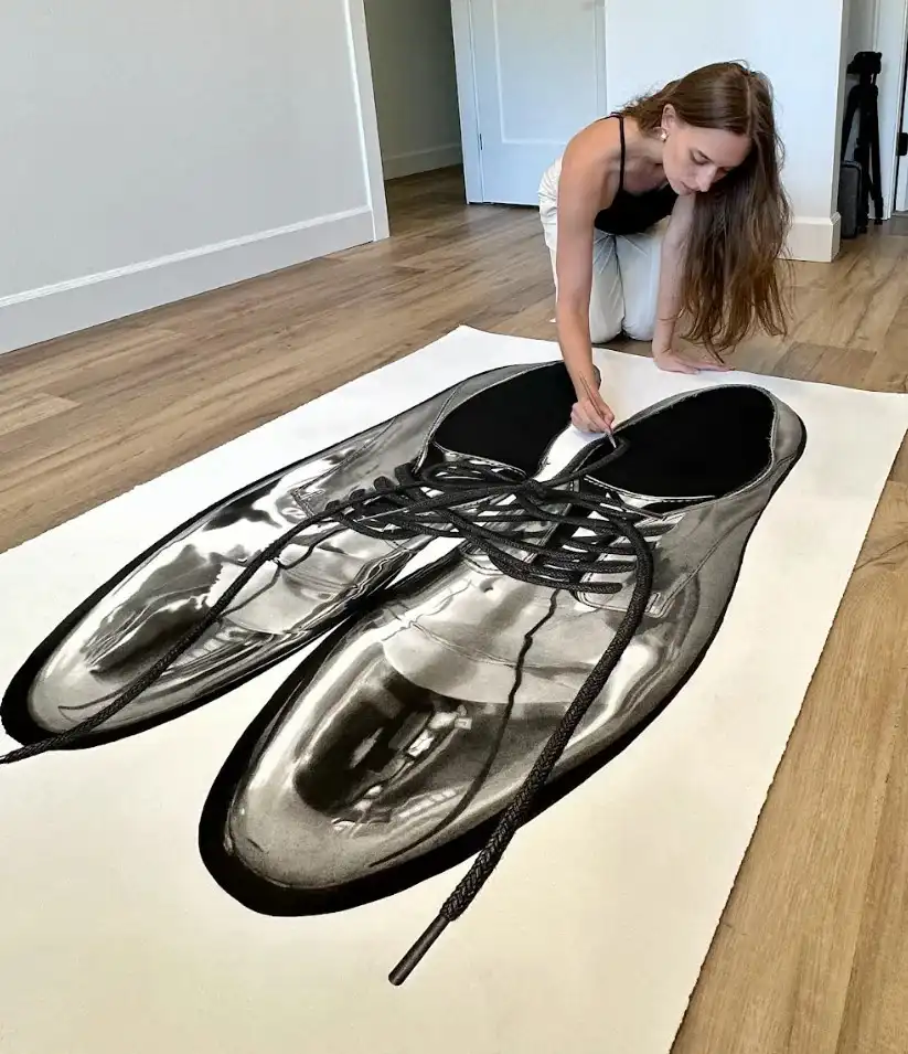 A woman kneels on a wooden floor beside a large, hyperrealistic black-and-white drawing of shiny dress shoes on a white rug…