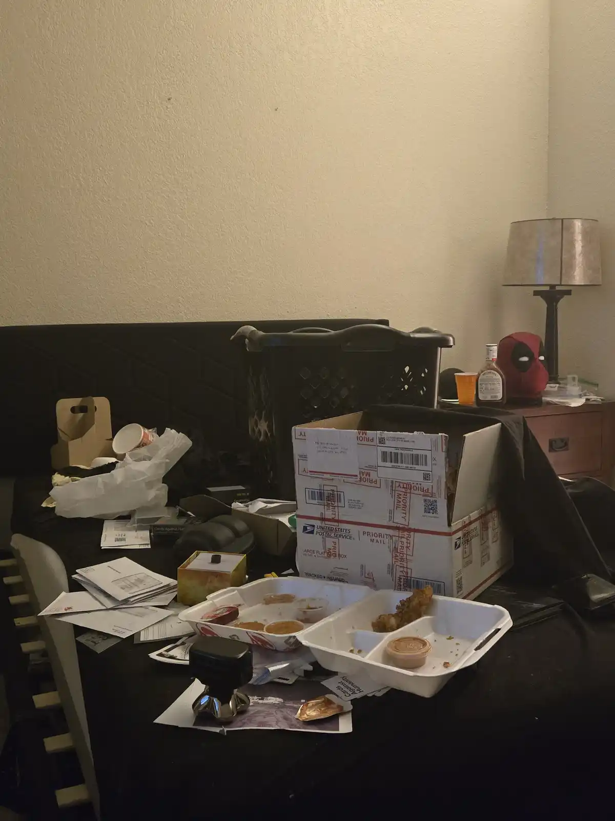 A messy room with a black couch and table covered in trash, takeout containers, papers, and a USPS Priority Mail box. A…