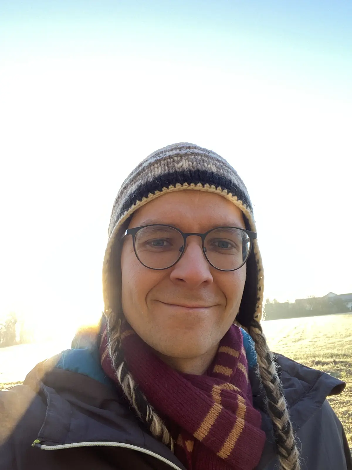 A man wearing glasses, a knitted winter hat with braided ear flaps, and a maroon and gold striped scarf smiles at the camera…