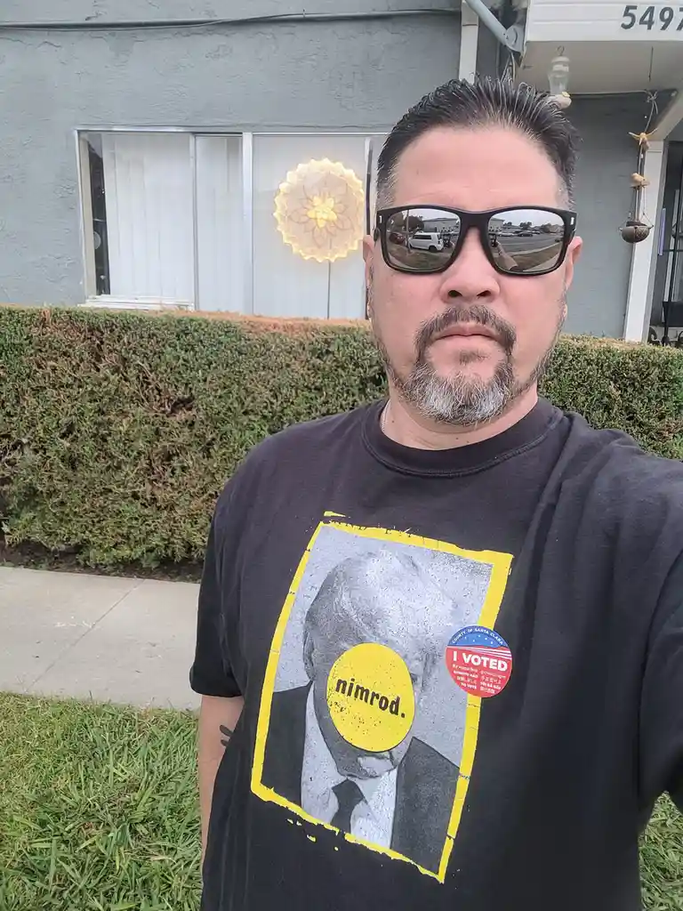 A man wearing sunglasses and a black t-shirt stands outside a house, the shirt features a graphic of a man's face with a…