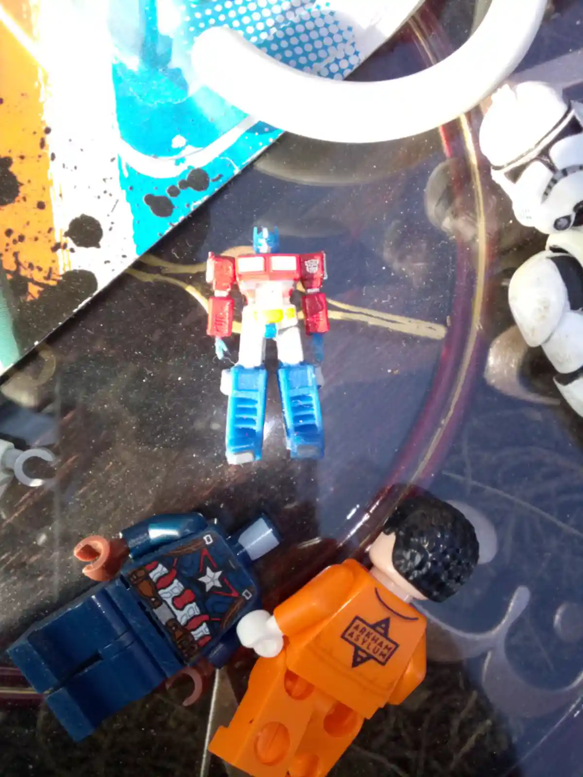 A small Optimus Prime action figure stands upright on a dark, reflective surface, surrounded by other toy figures including…