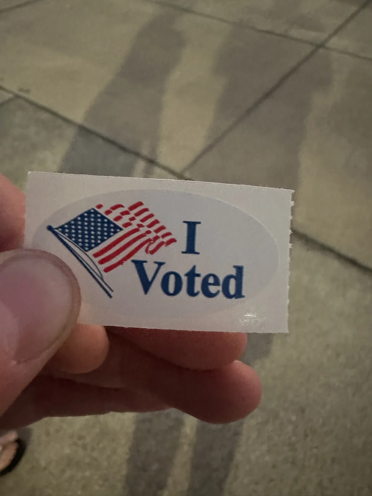 A close-up photo shows a person's hand holding a white oval sticker with a stylized American flag and the text 'I Voted' in…