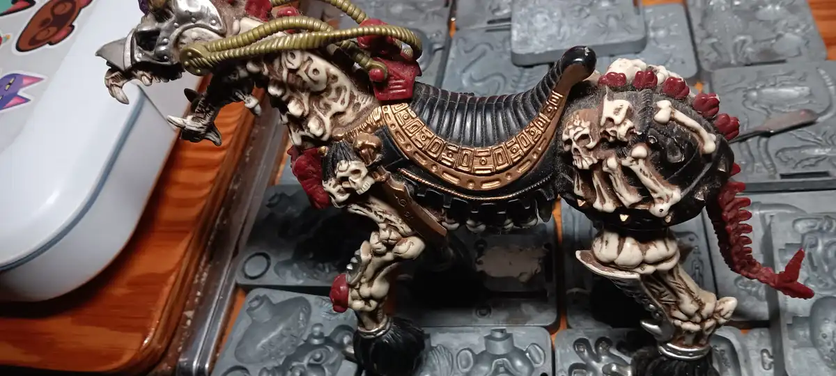 A highly detailed, gory miniature of a skeletal, demonic creature with skulls, bones, and red accents stands on a grey…
