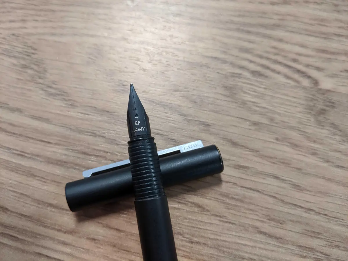A close-up photo shows a black Lamy fountain pen with its cap resting beside it on a wooden surface. The pen's nib is…