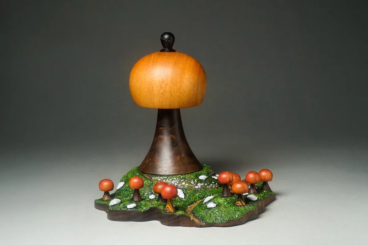 A wooden sculpture depicts a large mushroom with a smaller mushroom cluster around its base on a grassy, rocky terrain. The…