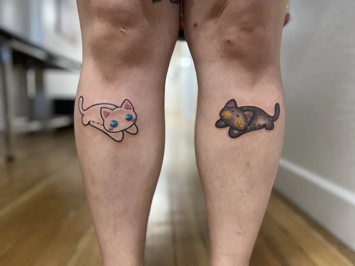 A person shows two matching cat tattoos on their knees, one on each leg. The left tattoo is a simple, outlined, pink cat…