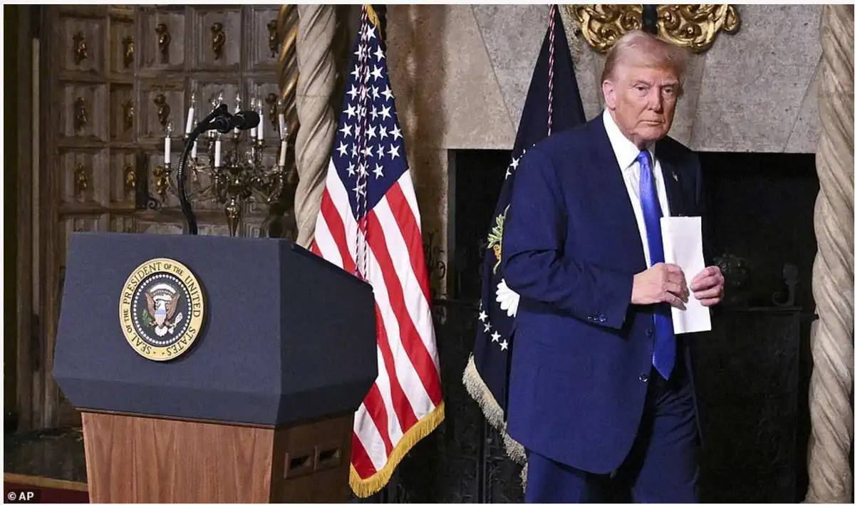 a man in a blue suit and blue tie stands beside a podium with the Seal of the President of the United States, holding a…