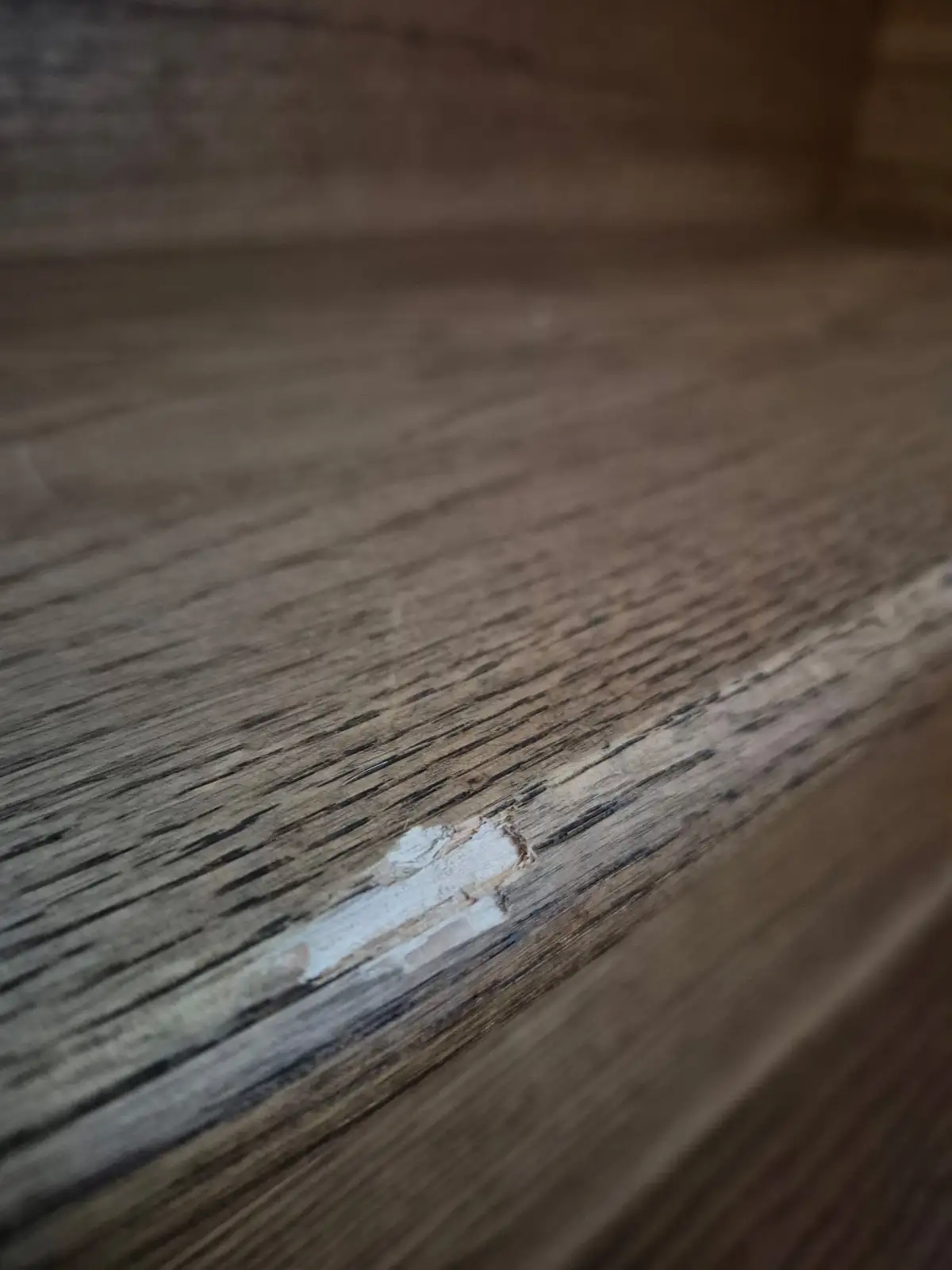 A close-up photo of a wooden surface with a small, noticeable chip or scratch revealing lighter wood underneath, overlaid…