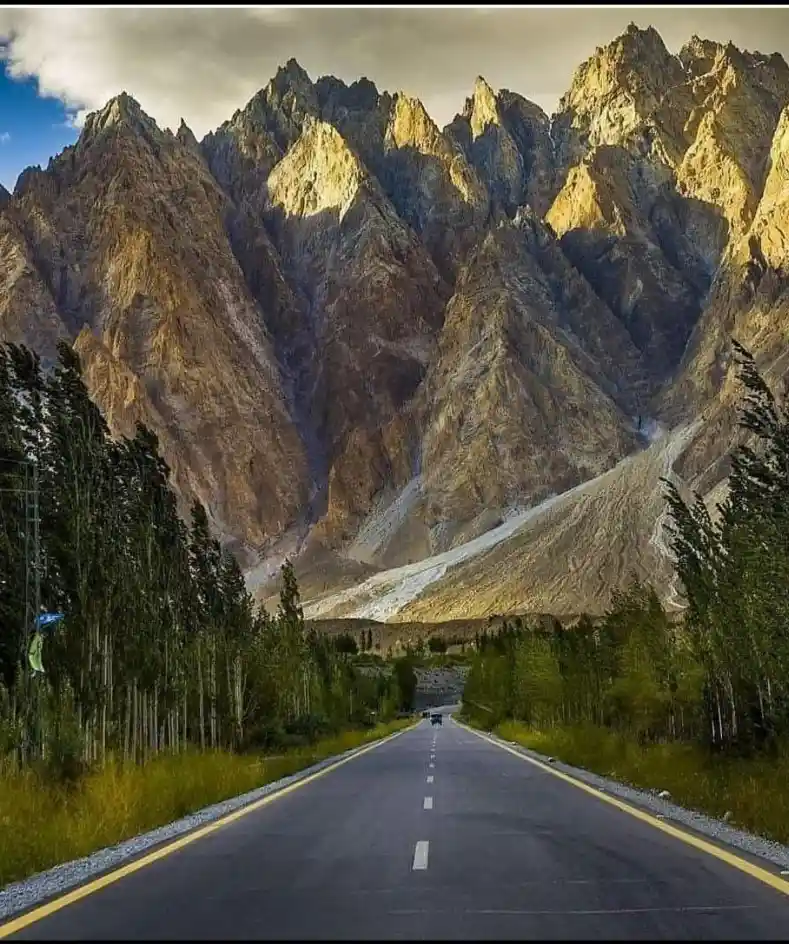 A scenic photo shows a long, straight asphalt road stretching toward distant, towering, jagged mountains under a partly…