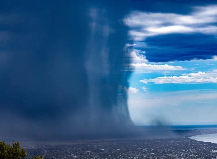 A dramatic landscape photo shows a massive, dark storm cloud descending over a city, contrasting sharply with a bright blue…