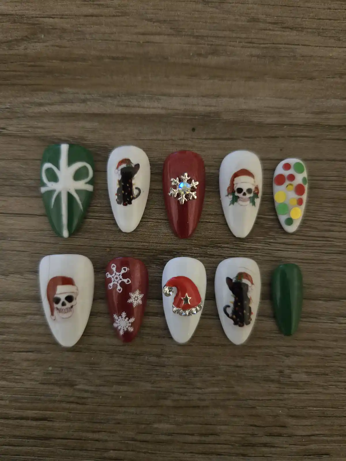 Ten artificial fingernails arranged in two rows on a wooden surface, decorated with various Christmas-themed designs…