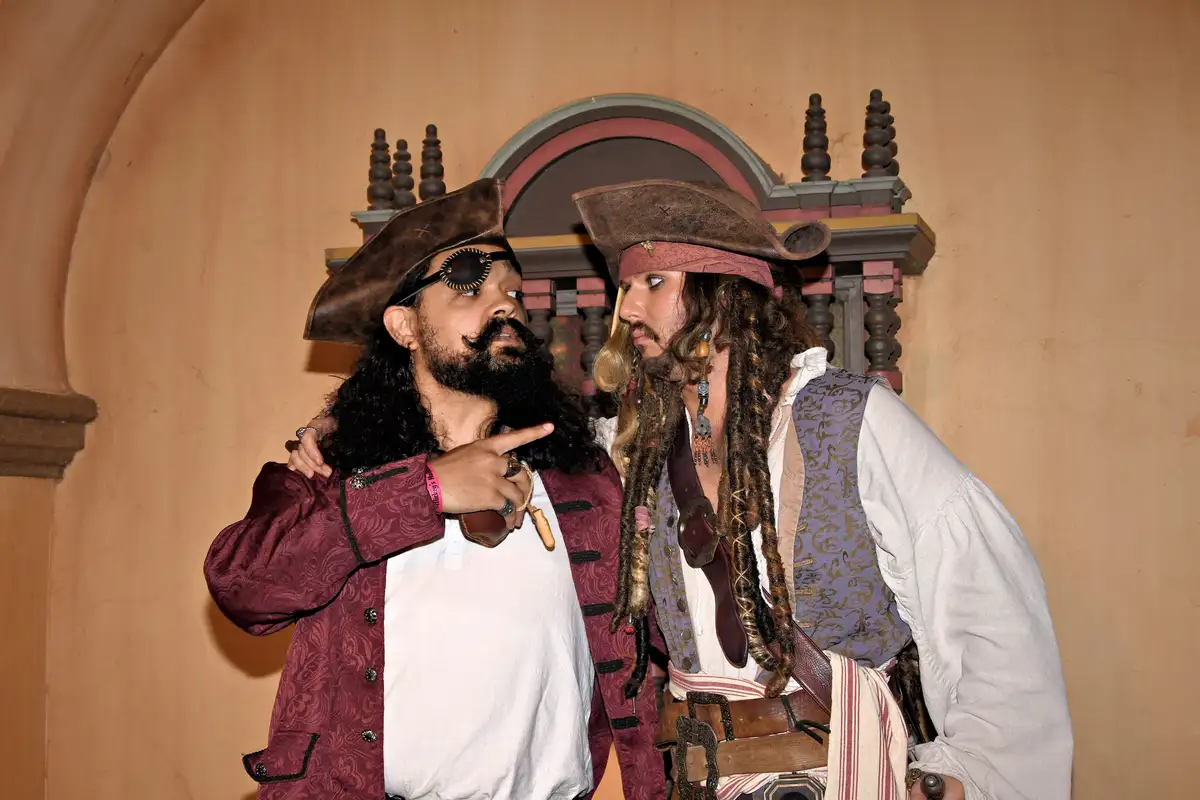 Two men dressed as pirates stand close together; the man on the left, wearing an eyepatch and a maroon coat, points at the…