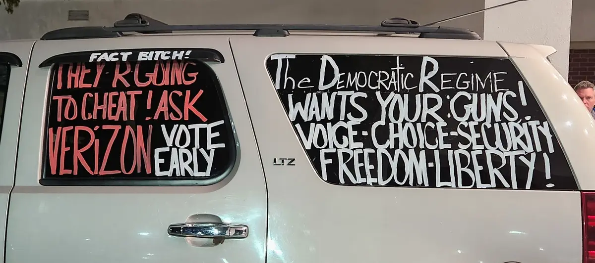 The rear windows of a white SUV are covered in handwritten political slogans. The left window reads 'FACT BITCH! THEY R…