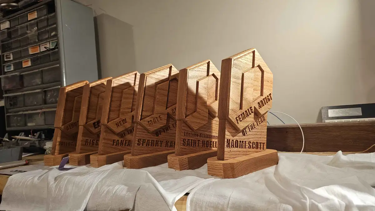 A row of handmade wooden award trophies sits on a white cloth, each engraved with different music award categories like…