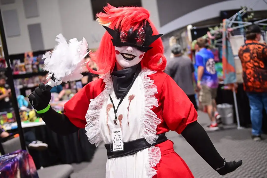 A person in a cosplay costume featuring a red wig, black mask, and a white dress with red stains, holding a white feather…