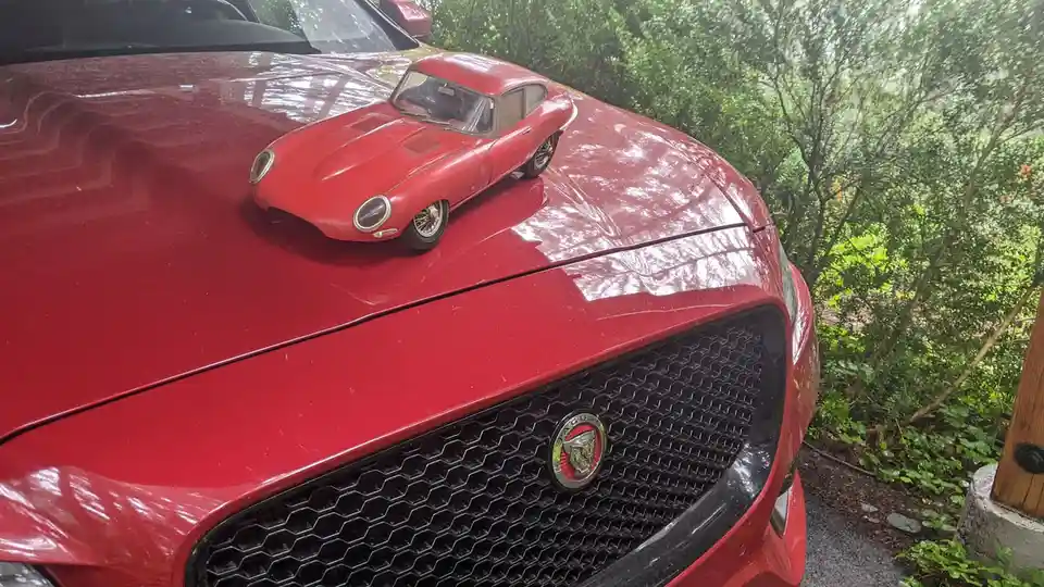 A red modern Jaguar car has a smaller red vintage Jaguar toy car placed on its hood. The toy car is a classic model, and the…