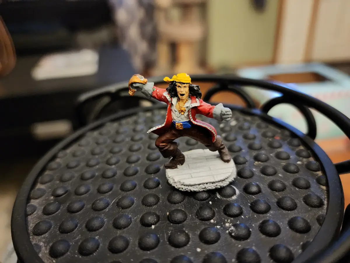 A painted miniature pirate figurine with a yellow bandana, red coat, and brown pants stands on a small white base atop a…