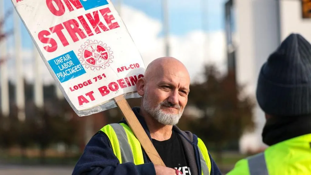 A bald man with a gray beard wearing a yellow safety vest holds a protest sign that reads 'ON STRIKE', 'UNFAIR LABOR…