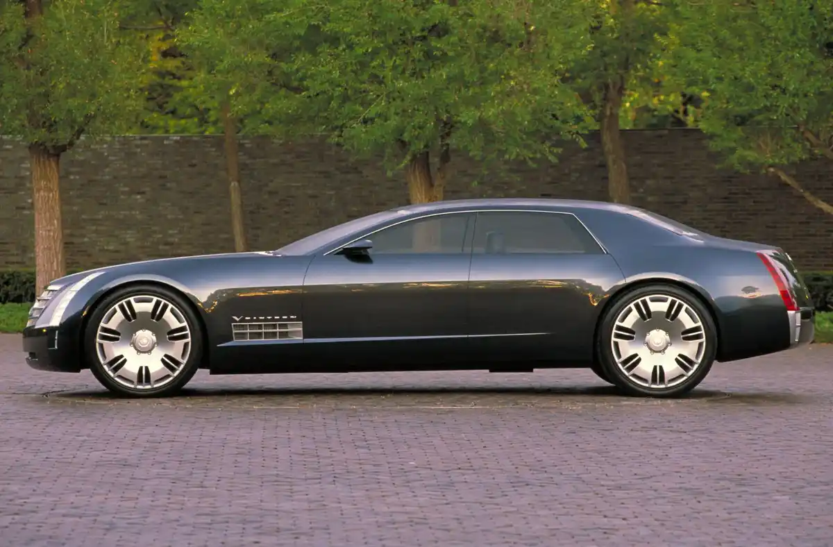 A sleek, dark gray Cadillac Sixteen concept car is shown in profile, parked on a cobblestone surface with trees and a brick…