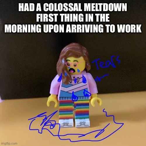 A Lego minifigure with brown hair, a distressed expression, and blue tears drawn on its face stands on a wooden surface…