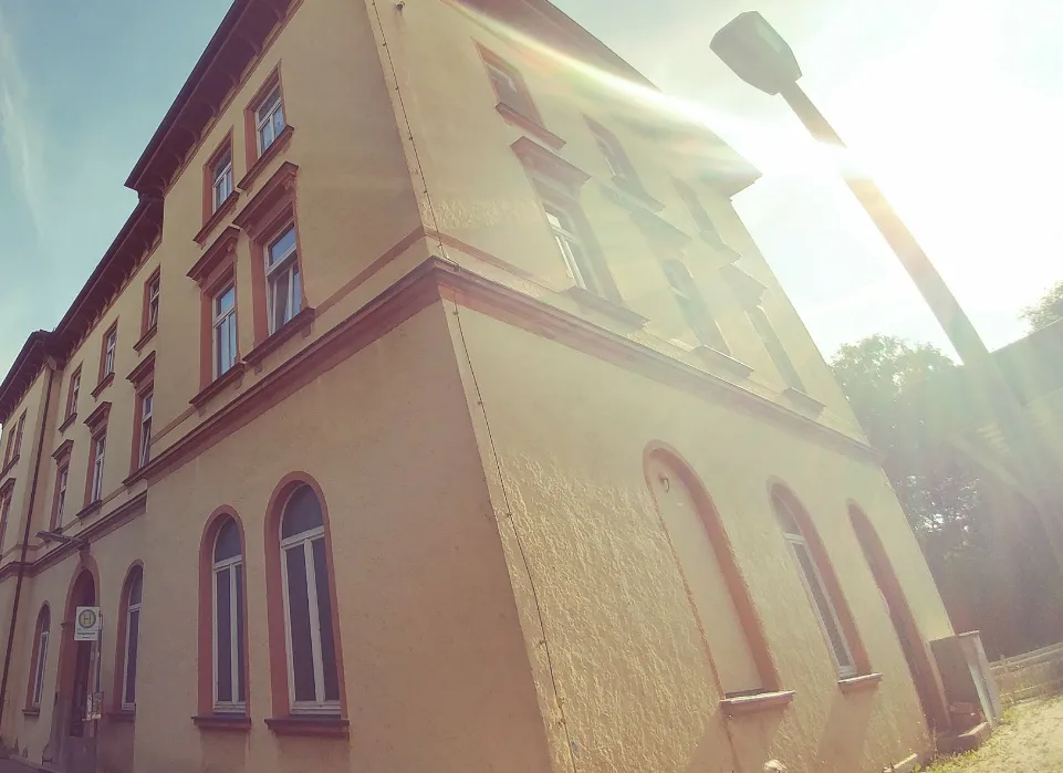 A low-angle photo of a beige, multi-story building with arched and rectangular windows, bathed in bright sunlight causing…