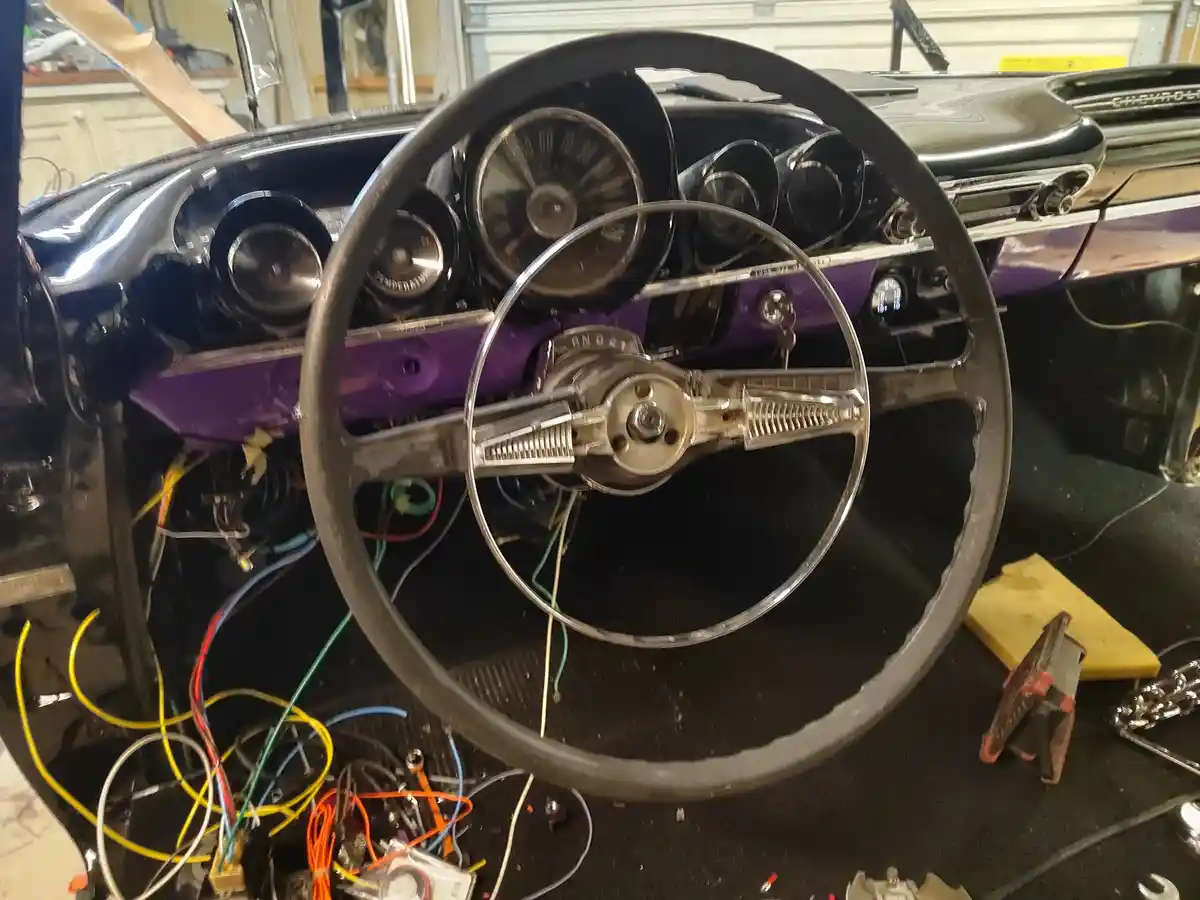 A close-up of a classic car's interior shows a steering wheel and dashboard with exposed wiring and tools scattered around…