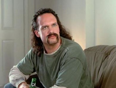 a man with long dark hair and a mustache, wearing a green t-shirt over a white long-sleeve shirt, sits on a couch holding a…