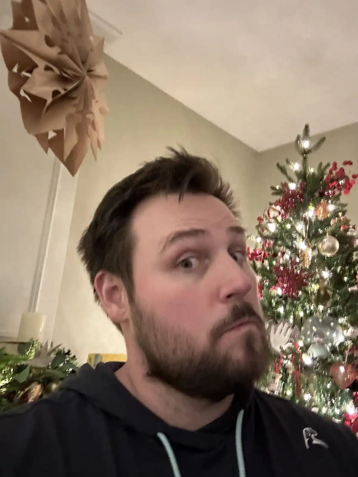 A man with a beard and dark hoodie looks upward with a deadpan, slightly disappointed expression. Behind him is a brightly…