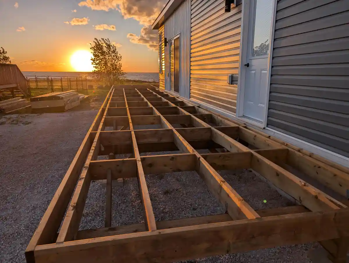 A wooden deck frame is being built on gravel next to a house, with a beautiful sunset over the ocean in the background. The…