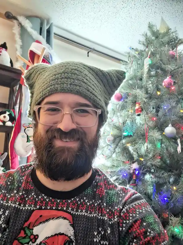 A smiling man with a beard and glasses wears a green knitted hat with cat ears and a festive sweater with a Santa design…