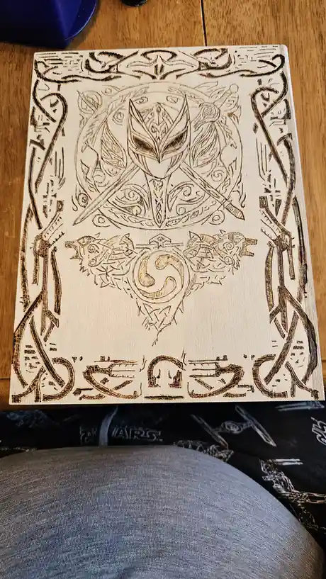 A wooden board features a detailed pyrography design of a stylized mask with crossed swords, flanked by wolves and ornate…