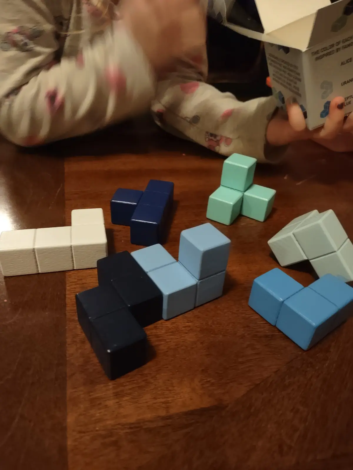 A child in a patterned pajama top is playing with colorful, cube-shaped blocks on a wooden floor, arranging them into…