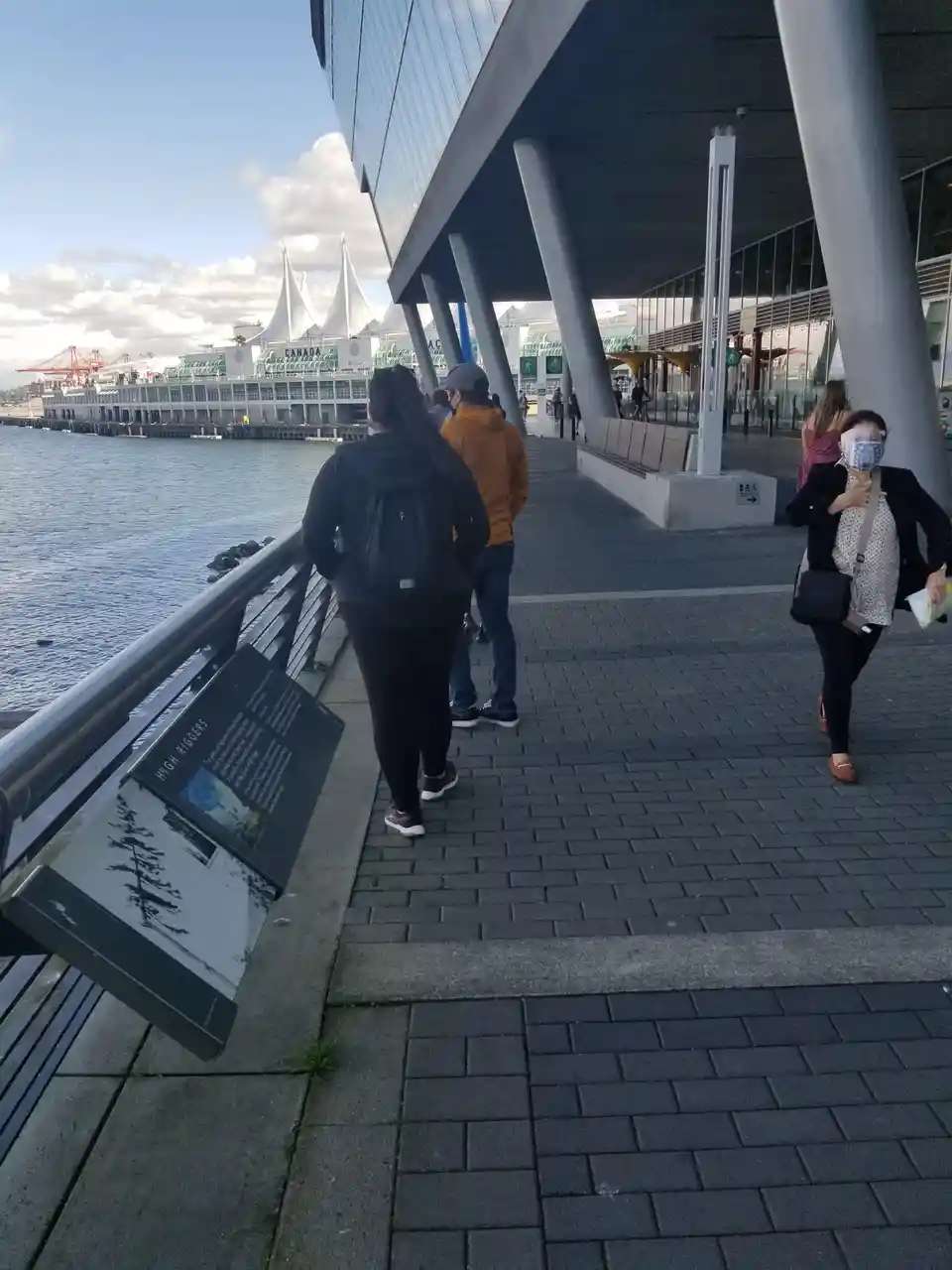 A tilted informational sign on a waterfront promenade reads 'HIGH RIGGERS' with a tree illustration, while pedestrians walk…