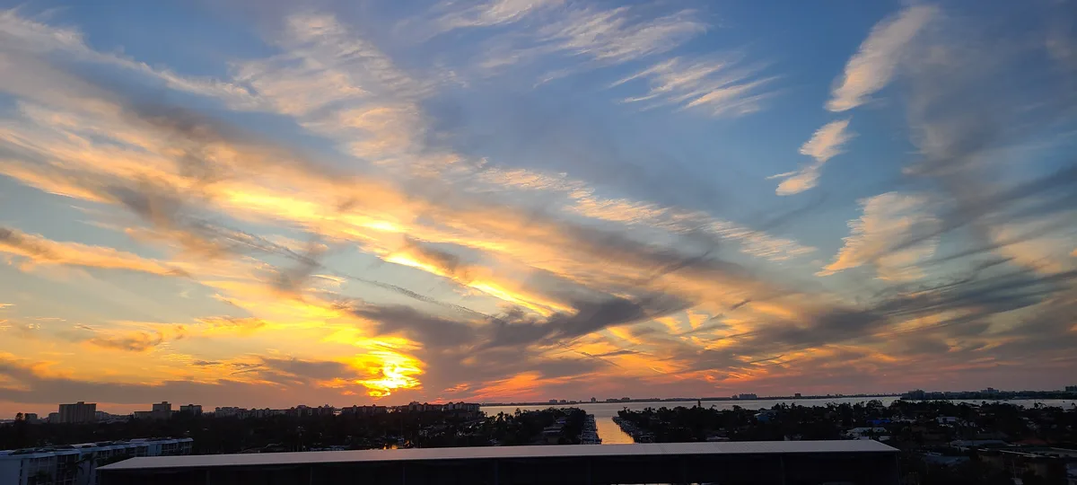 A panoramic photo of a vibrant sunset over a cityscape and water, with streaks of orange, yellow, and blue clouds. The sun…
