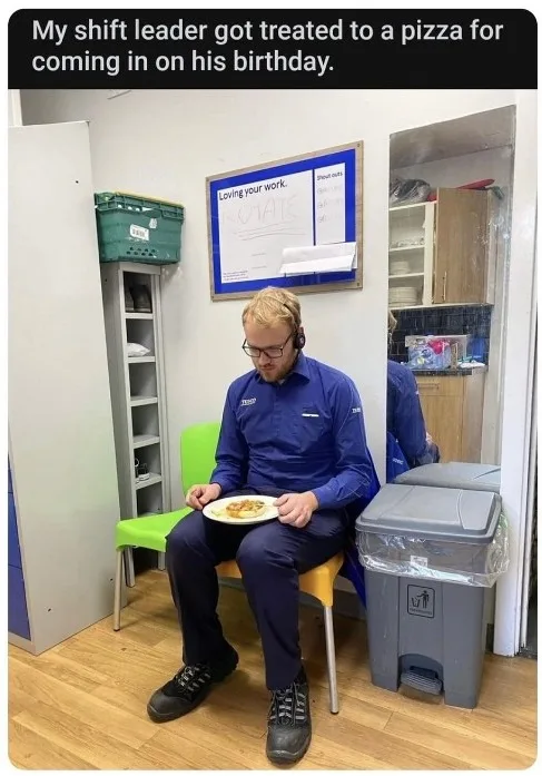 A man in a blue work uniform sits on a green and yellow chair in a breakroom, holding a plate with a small, plain-looking…