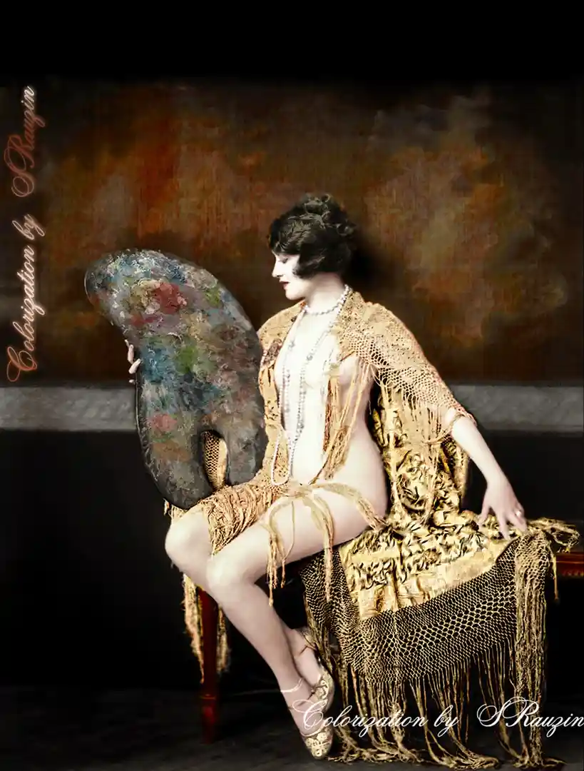 A colorized vintage photograph shows a woman in a gold fringed robe sitting on a chair, holding a painter's palette. She is…