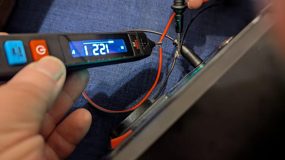 A close-up shows a hand holding a black multimeter with a blue LCD screen displaying the number '1221' and an ampere symbol…