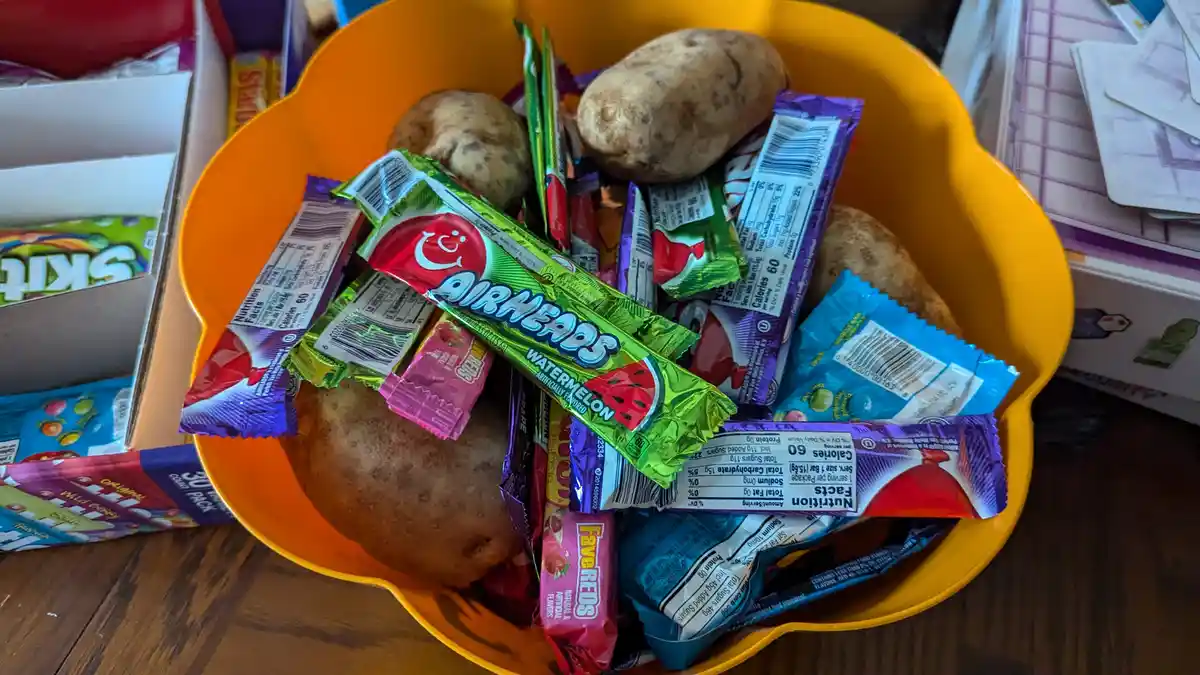 An orange bowl filled with various individually wrapped candies like Airheads and Fave Reds, mixed with several whole…