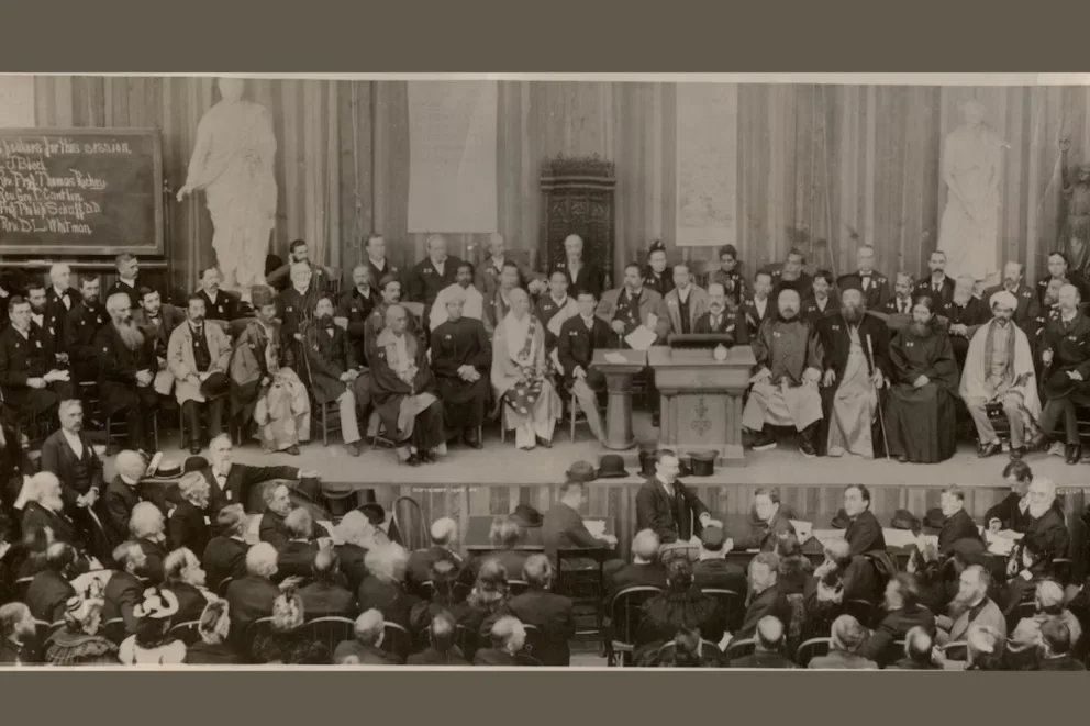 A sepia-toned historical photograph shows a large gathering of men in formal attire seated in rows on a stage and in an…