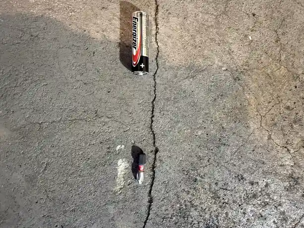 An Energizer battery and a small screwdriver lie on cracked concrete, positioned on opposite sides of a deep crack. The…