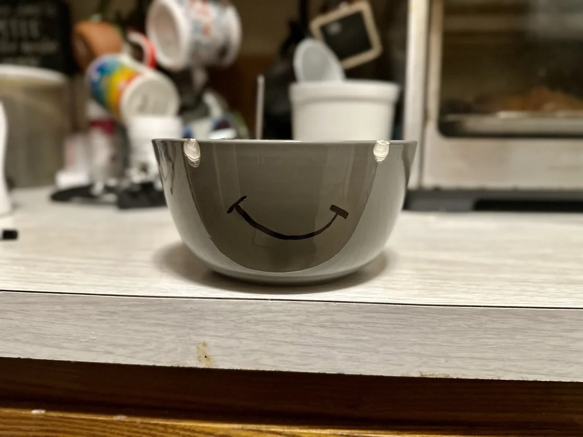 A gray ceramic bowl with a hand-drawn smiley face on its side sits on a white countertop, visibly chipped on both sides of…