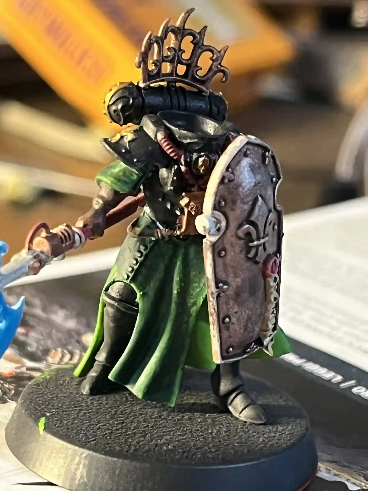 A painted miniature of a heavily armored warrior with a green cloak, holding a sword and shield, wears a decorative crown on…