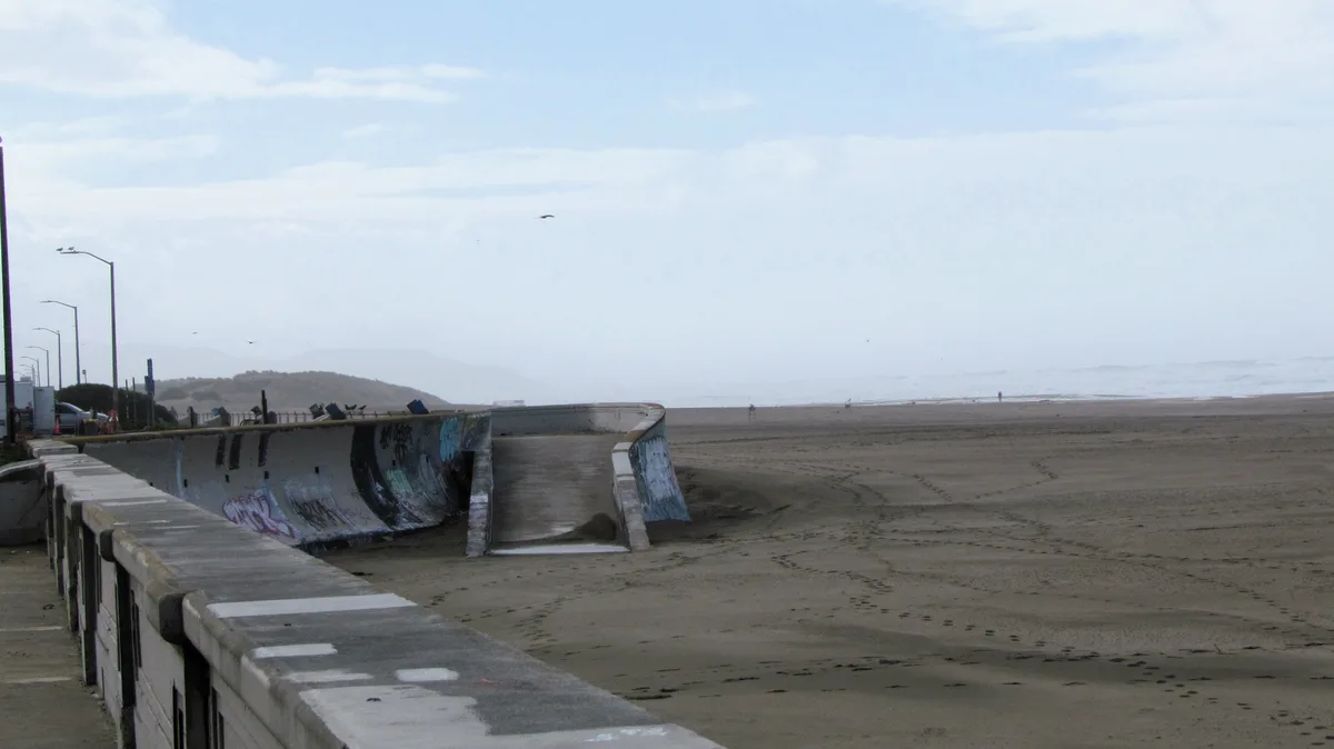A wide shot of a desolate beach with a large, graffiti-covered concrete structure resembling a half-pipe or skate ramp…